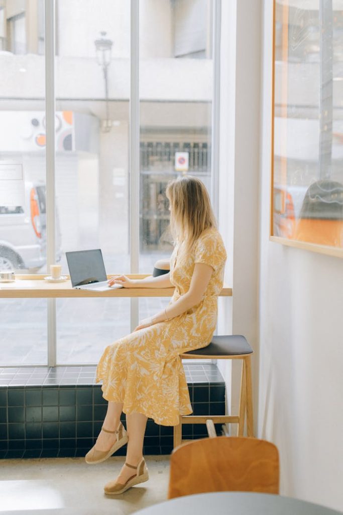 Woman in a yellow dress working on a laptop by a window in a cafe, embracing remote work lifestyle.